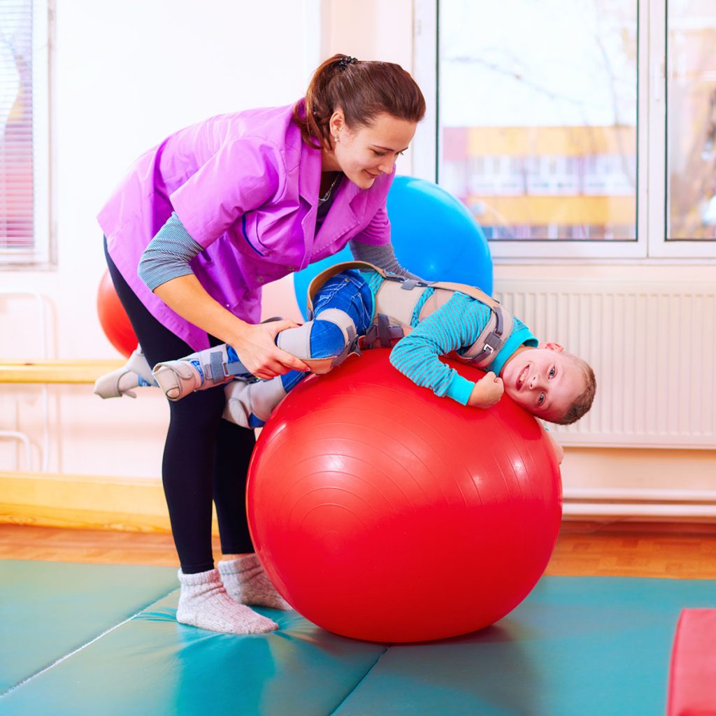 Cute kid with disability has musculoskeletal therapy by doing exercises in body fixing belts on fit ball